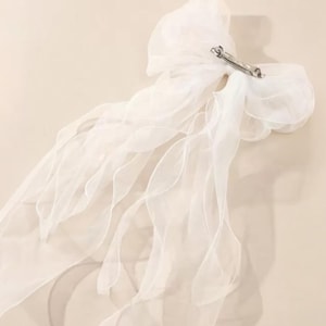 Op de afbeelding: Een witte, doorschijnende stoffen haarstrik met een zilverkleurige metalen clip. De strik heeft meerdere lagen vloeiende stof, waardoor een delicate en luchtige uitstraling ontstaat. Het accessoire is ontworpen voor haarstyling.