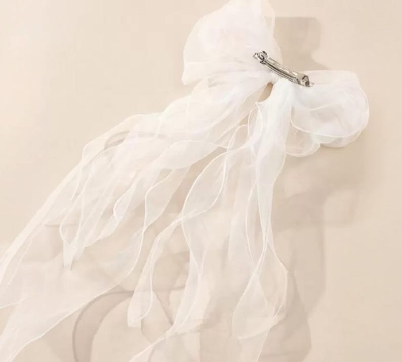 Op de afbeelding: Een witte, doorschijnende stoffen haarstrik met een zilverkleurige metalen clip. De strik heeft meerdere lagen vloeiende stof, waardoor een delicate en luchtige uitstraling ontstaat. Het accessoire is ontworpen voor haarstyling.