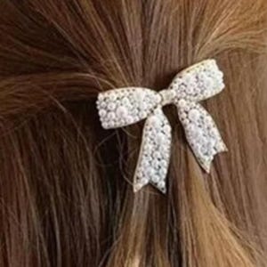 Puede incluir: Una pinza para el pelo con forma de lazo blanco, adornada con perlas, est&aacute; sujeta en cabello casta&ntilde;o. El lazo presenta un dise&ntilde;o intrincado con una superficie delicada y texturizada, creando un efecto brillante. La pinza a&ntilde;ade un toque de elegancia a cualquier peinado.