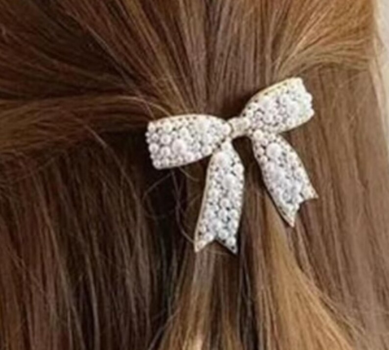 Puede incluir: Una pinza para el pelo con forma de lazo blanco, adornada con perlas, est&aacute; sujeta en cabello casta&ntilde;o. El lazo presenta un dise&ntilde;o intrincado con una superficie delicada y texturizada, creando un efecto brillante. La pinza a&ntilde;ade un toque de elegancia a cualquier peinado.