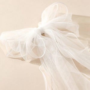 Op de afbeelding: Een close-up van een witte tule strik, waarschijnlijk voor een bruidsaccessoire. De strik is volumineus en luchtig, met meerdere lagen transparante stof. De achtergrond is een zachte, neutrale tint, die de fijne details en textuur van de strik benadrukt.