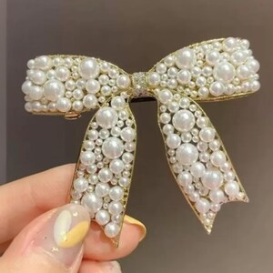 Puede incluir: Pinza para el cabello dorada con forma de lazo, adornada con numerosas perlas de imitaci&oacute;n. Las perlas, de diferentes tama&ntilde;os, crean una textura rica. El accesorio est&aacute; dise&ntilde;ado para sujetar el cabello y a&ntilde;ade elegancia.