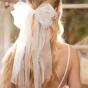 Op de afbeelding: Een witte, doorschijnende stoffen strik en sluier zijn bevestigd aan blond haar. De strik is groot en volumineus, met meerdere lagen stof. De sluier valt over de rug en voegt een vleugje elegantie toe. De algehele uitstraling is zacht en romantisch.