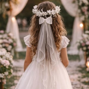 May include: A young person in a white dress with a floral crown and a veil. The veil is adorned with a white bow and a string of pearls. The dress has lace sleeves. The background is blurred, suggesting an outdoor event.