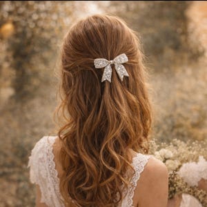 Puede incluir: Una mujer con cabello largo y ondulado de color casta&ntilde;o rojizo lleva un pasador para el pelo con forma de lazo adornado con brillantes pedrer&iacute;a. El pasador est&aacute; en el centro del cabello. La mujer lleva un vestido de encaje blanco. El fondo est&aacute; borroso.