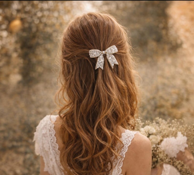 Puede incluir: Una mujer con cabello largo y ondulado de color casta&ntilde;o rojizo lleva un pasador para el pelo con forma de lazo adornado con brillantes pedrer&iacute;a. El pasador est&aacute; en el centro del cabello. La mujer lleva un vestido de encaje blanco. El fondo est&aacute; borroso.
