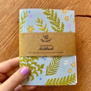 May include: A light blue notebook featuring a botanical pattern of green ferns and small yellow and pink flowers. A brown paper band wraps around the centre, printed with the text Paper Moth co. HANDMADE Notebook FILLED WITH 30 UNLINED PAGES.