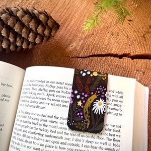 Può includere: Un segnalibro magnetico rettangolare con sfondo scuro che raffigura una lumaca gialla, margherite bianche, piccoli fiori rosa e pigne marroni. Il segnalibro è inserito in un libro aperto con testo stampato visibile sulle pagine.