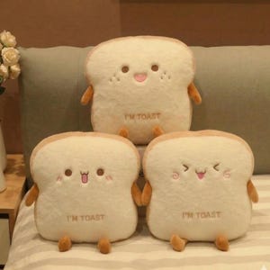 May include: Three plush bread slice pillows with smiling faces, arms, and legs. The pillows are off-white with tan edges, and the text "I'M TOAST" is printed on each. The pillows are on a bed with a green pillow in the background.