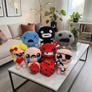 Op de afbeelding: Een verzameling pluche speelgoed met verschillende stripfiguren met verschillende gezichtsuitdrukkingen en kleuren. De knuffels zijn gerangschikt op een witte marmeren tafel, samen met een grote rode dobbelsteen en een zwart-wit knuffel.