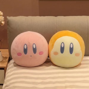 Peut inclure: Deux coussins ronds en peluche. L'un est rose avec des yeux ovales bleus et des joues roses. L'autre est jaune avec un visage blanc, des yeux ovales bleus et des joues roses. Les deux coussins sont sur une couverture à rayures.