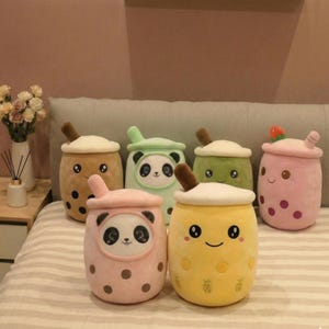 Puede incluir: Una colección de cojines de peluche con forma de té de burbujas en varios colores, incluyendo marrón, verde, rosa y amarillo. Cada cojín presenta una cara sonriente y una pajita marrón. Algunos cojines tienen detalles de panda o fresa.
