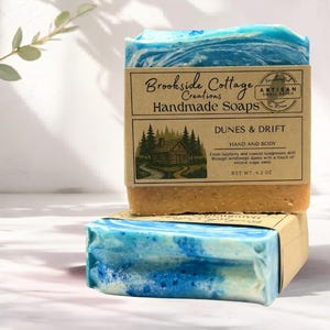 May include: Two bars of handmade soap with a blue and white marbled design, labelled "Dunes & Drift." The soap has a tan base and is wrapped in a brown paper label with the text "Brookside Cottage Creations Handmade Soaps."