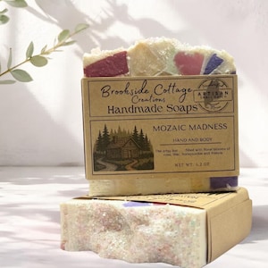Puede incluir: Barras de jabón hechas a mano de Brookside Cottage Creations. Los jabones están en una caja con el texto "MOZAIC MADNESS" y "HAND AND BODY". El jabón está relleno de flores de rosa, lila, madreselva y fresia. Peso neto: 119 gramos.