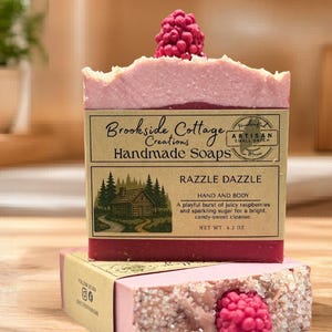 May include: A handmade Razzle Dazzle soap bar with a raspberry-shaped top, resting on a box of soap. The soap is pink with a textured top and a label that reads "Brookside Cottage Creations Handmade Soaps". The net weight is 119 grams.