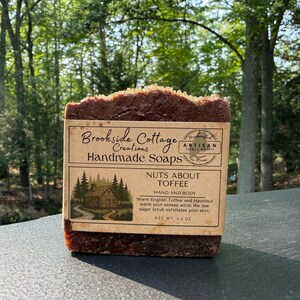 Könnte beinhalten: Ein rechteckiges Stück handgemachte Seife in Dunkelbraun mit strukturierter Oberfläche, umwickelt mit einem beigen Papieretikett. Das Etikett zeigt den Text Brookside Cottage Creations Handmade Soaps, Nuts About Toffee und eine Waldhütte.