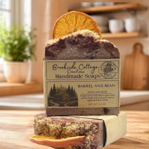 May include: A handmade soap bar from Brookside Cottage Creations, with a slice of dried orange and sugar crystals on top. The label reads "Barrel and Bean" and "Hand and Body". The soap weighs 4.2 oz.