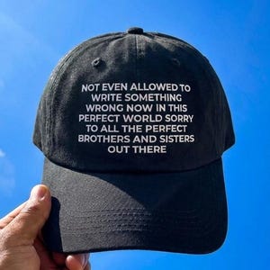 May include: A black baseball cap featuring white embroidered text that reads: NOT EVEN ALLOWED TO WRITE SOMETHING WRONG NOW IN THIS PERFECT WORLD SORRY TO ALL THE PERFECT BROTHERS AND SISTERS OUT THERE. The cap has a curved brim and a structured crown.
