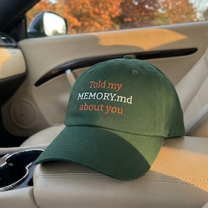 Peut inclure: Une casquette de baseball vert forêt avec du texte brodé sur le panneau avant. Le texte indique "Told my MEMORY.md about you", avec les mots "Told my" et "about you" en orange, et "MEMORY.md" en blanc, centrés sur la calotte.