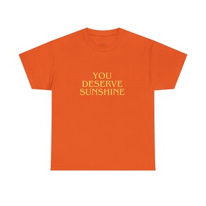 May include: An orange t-shirt with the gold text "YOU DESERVE SUNSHINE". The t-shirt has a crew neck and is a solid orange colour.