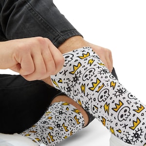 May include: White socks with a repeating pattern of black skulls, yellow crowns, lightning bolts, and other doodles. The socks are being pulled up, showcasing the design.