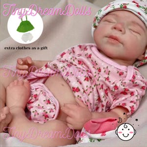 May include: A lifelike baby doll dressed in a pink floral print onesie and matching hat. The doll has closed eyes and a pacifier. The image includes a graphic of extra clothes as a gift.