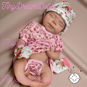May include: A realistic baby doll dressed in a pink floral print ensemble, comprising a hat, shirt, nappy, and mittens. The doll has closed eyes and is lying down. The text "Tiny Dream Dolls" is visible in the image.
