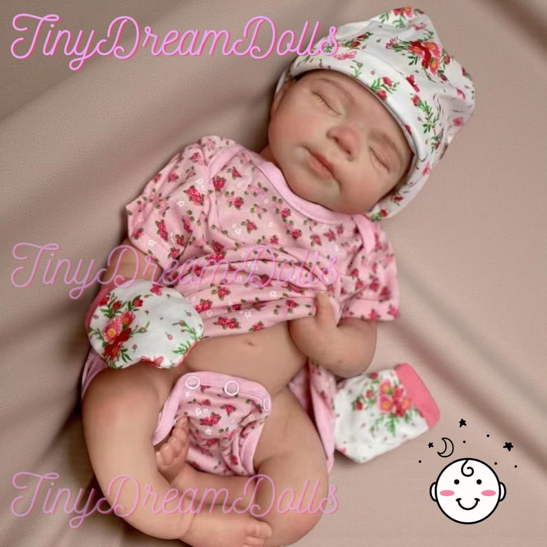 May include: A realistic baby doll dressed in a pink floral print ensemble, comprising a hat, shirt, nappy, and mittens. The doll has closed eyes and is lying down. The text "Tiny Dream Dolls" is visible in the image.