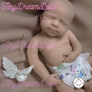 May include: A realistic baby doll with closed eyes and a natural skin tone. The doll is wearing a diaper with a playful design. A small, white, winged figurine with a gold wire is next to the doll. The text "Tiny Dream Dolls" is in pink cursive.