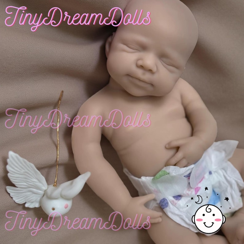 May include: A realistic baby doll with closed eyes and a natural skin tone. The doll is wearing a diaper with a playful design. A small, white, winged figurine with a gold wire is next to the doll. The text "Tiny Dream Dolls" is in pink cursive.