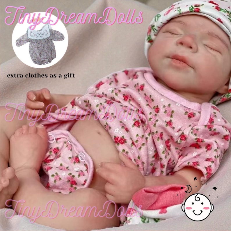 May include: A realistic baby doll dressed in a pink floral onesie and matching nappy cover. The doll has a white floral hat and is lying on a soft surface. The image includes the text "Tiny Dream Dolls" and "extra clothes as a gift".