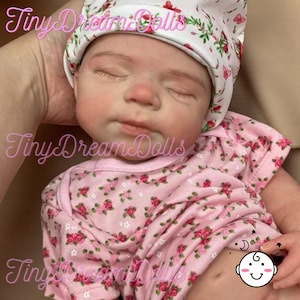 May include: A realistic baby doll with closed eyes, dressed in a pink floral print onesie and a matching hat. The doll has a lifelike skin tone and delicate facial features. The doll is held in someone's hands.