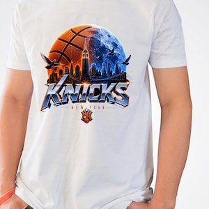 May include: A white short-sleeved t-shirt featuring a graphic of a basketball merged with a moon over the New York City skyline. The text KNICKS appears in large blue metallic letters above the words NEW YORK and a small orange basketball logo.