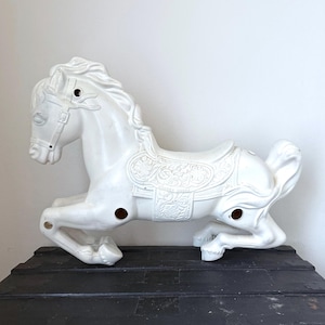 Puede incluir: Un caballo balancín blanco de estilo vintage, con silla de montar y brida detalladas. El caballo está hecho de un material sólido y tiene un diseño clásico. El caballo está colocado sobre un baúl negro.