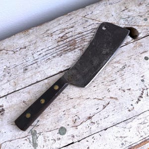 May include: A vintage cleaver with a dark, aged blade and a wooden handle. The handle is secured with brass rivets. The cleaver has a hole near the top of the blade. The cleaver is resting on a distressed, white-painted wooden surface.