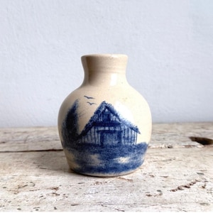 May include: A small, cream-colored ceramic vase with a rounded body and a narrow neck. The surface features a blue painted illustration of a house with a gabled roof, surrounded by trees and two small birds flying in the sky above the structure.