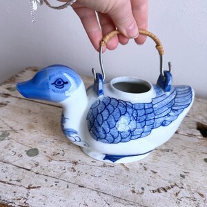 May include: A ceramic duck-shaped teapot in white and blue. The teapot has a blue beak, eye, and wing details. It features a woven handle and metal supports. The teapot is on a distressed wooden surface.