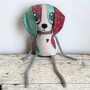May include: A stuffed dog toy made of beige fabric with a patchwork design. It features one green ear and one red floral patterned ear, large black eyes, a brown collar with a small metal ring, and long gray cord limbs. A small red heart is stitched on the chest.