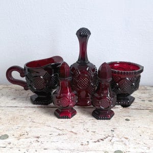 Könnte beinhalten: Ein Set aus dunkelrotem Pressglas-Geschirr, bestehend aus einem Milchkännchen, einer Zuckerdose, einer hohen Karaffe und zwei Streuern. Jedes Teil weist eine gleichmäßige Rautenstruktur mit kreisförmigen Akzenten in einer satten, rubinroten Farbe auf.