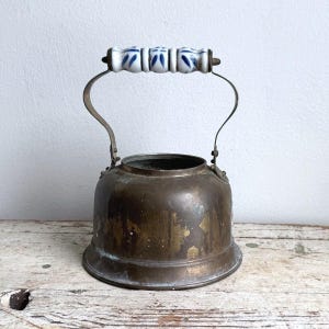 May include: A vintage brass kettle with a weathered, tarnished patina and a wide, open top. The kettle features a tall, arched metal handle topped with a cylindrical white ceramic grip decorated with a repeating blue floral pattern.