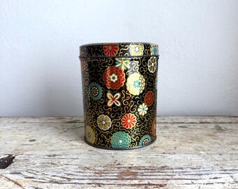 May include: A cylindrical black metal tin with a lid, adorned with a colorful floral pattern. The flowers are in shades of red, yellow, blue, and white, set against a background of gold swirls. The tin is likely for storage.
