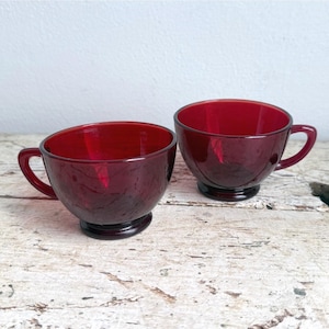 Può includere: Due tazze da tè abbinate in vetro rosso rubino con una finitura liscia e traslucida. Ogni tazza presenta una forma a ciotola arrotondata, una base circolare robusta e un manico curvo sul lato, mostrando un colore cremisi profondo e vivace.