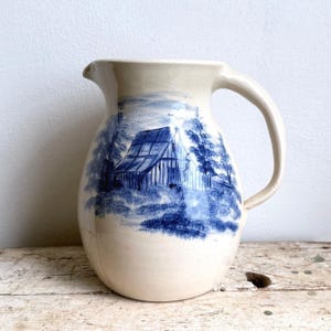Puede incluir: Una jarra de cerámica de color crema con una ilustración azul pintada a mano de un granero rodeado de árboles. La jarra tiene un cuerpo redondeado, un pico ancho y un asa curva en el lado derecho, con un acabado de esmalte craquelado.