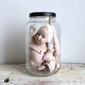 Op de afbeelding: Een doorzichtige glazen pot met een zwarte deksel die verschillende losse porseleinen poppenonderdelen bevat, waaronder een hoofd met geschilderde gelaatstrekken, meerdere armen en benen, allemaal in een rommelige opstelling.