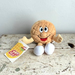 May include: A plush toy shaped like an Eggo waffle with a smiling face, arms, and legs. The waffle is tan with a quilted pattern. It has white eyes, a red mouth, and white feet. A tag with the Eggo logo is attached.