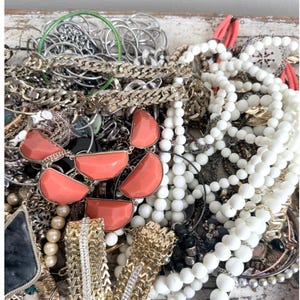 May include: A selection of jewellery items, including necklaces, bracelets, and earrings. The collection features gold and silver chains, coral-coloured pendants, white beaded necklaces, and other decorative elements. The jewellery is piled together, creating a textured and layered appearance.