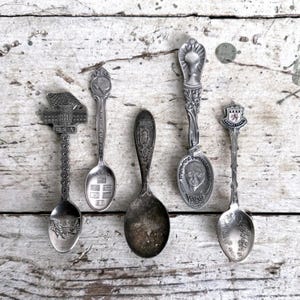 May include: Five antique silver spoons with intricate designs. One spoon features the text "USA" and an eagle, another has the Texas state outline, and a third has a portrait of Thomas Edison. The spoons are arranged on a distressed, white wooden surface.