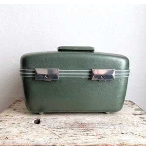 May include: A forest green hard-shell train case with a textured finish. The rectangular vanity case features a top handle, two silver-toned metal latches on the front, and three horizontal white stripes running across the center of the body.