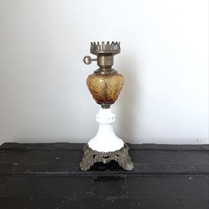 Pode incluir: Uma antiga lamparina a óleo com um design decorativo. A lamparina apresenta um reservatório de vidro transparente com um tom âmbar, uma base branca e um topo de metal. A base tem um pé de metal decorativo. É um artigo de decoração vintage.