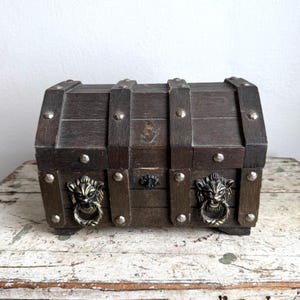 May include: A small, dark brown wooden chest with metal accents. The chest features decorative lion head handles and is reinforced with metal studs. The wood has a distressed appearance, suggesting an antique or vintage style.